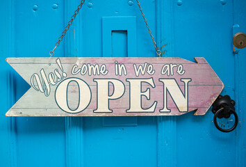 Close up of a wooden sign hanging by a chain saying "Yes!Come in we are open" against a blue door background in Lincoln, England.