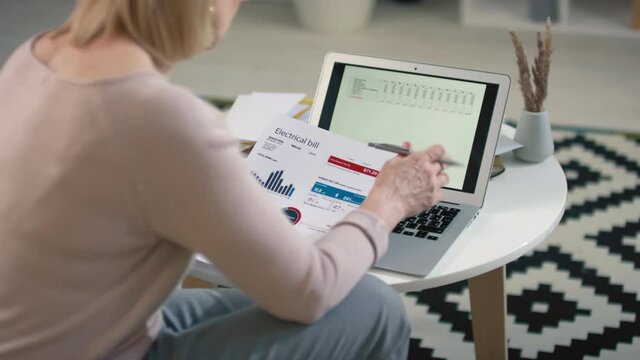 Back-view footage of caucasian woman sitting in front of laptop with opened planning spreadsheet managing budget according to monthly mandatory expenses looking at printed charts and diagrams