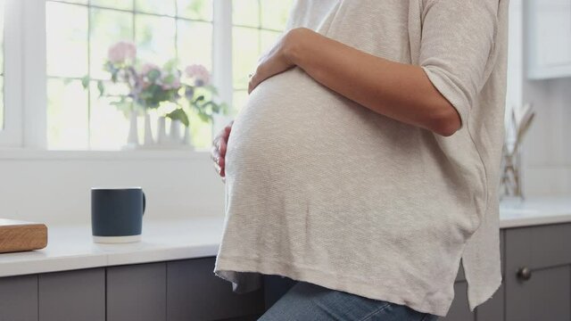 Close up of pregnant woman standing by kitchen window touching stomach relaxing with hot drink - shot in slow motion