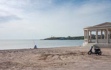 The sandy shore of the Azov sea with a fisherman, a scooter and a fragment of a gazebo in the...