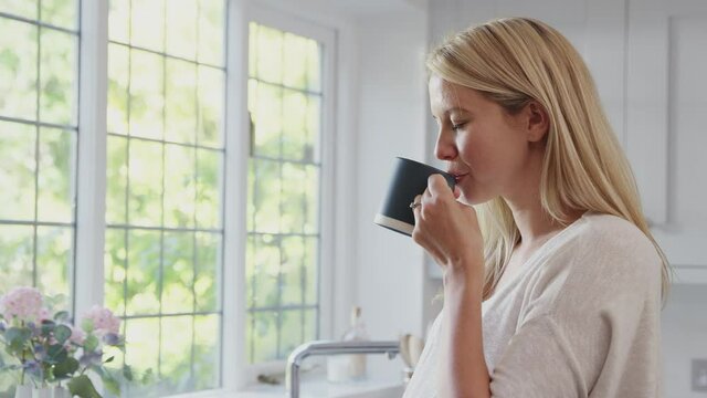Smiling pregnant woman standing by kitchen window touching stomach relaxing with hot drink - shot in slow motion