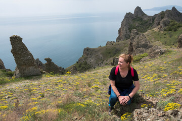 A female tourist on a slope with yellow flowers on the background of the Kara-Dag mountain range, the sea and the cloudy sky on the Crimean Peninsula