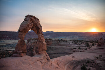 View of the delicate arch in arches national park at sunset. The sun can be seen and the land around it is lit up from the sunset. 