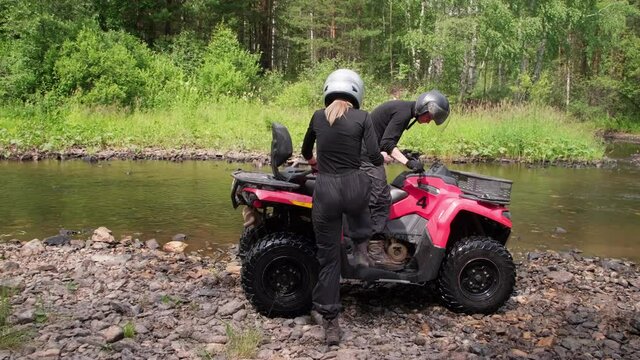 Tracking shot of man and woman in helmets getting on red quad bike and driving across shallow river in forest
