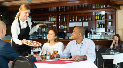 Portrait of young smiling waitress working in restaurant, serving ordered pizza to friendly company