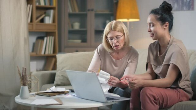 Middle aged caucasian woman learning financial accounting with help of younger daughter explaining how to pay bills and plan budget online using laptop sitting in living room