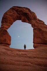 Person standing under Delicate Arch in Arches National Park during sunset. Person looks tiny compared to the famous rock formation.