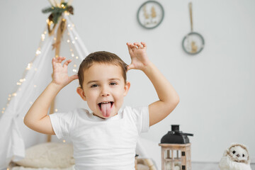Little Caucasian sticks out tongue and grimaces in Christmas Scandinavian interior.