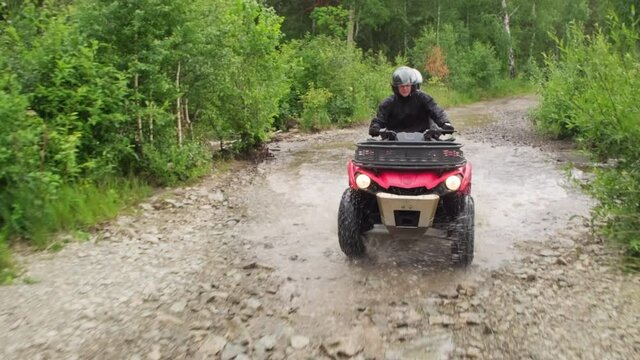 Tracking shot of man and woman in helmets driving red quad bike in rough terrain in forest
