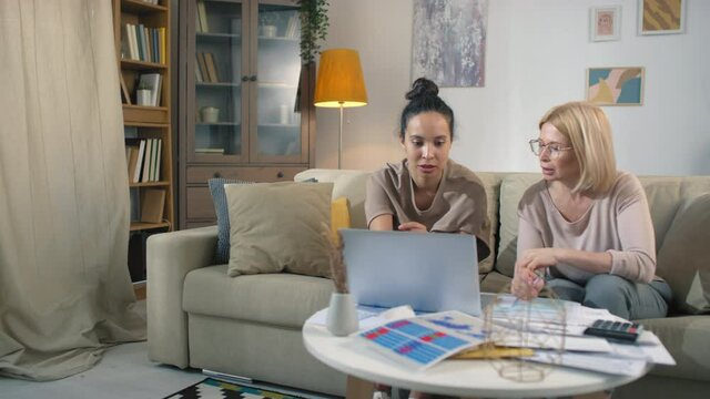 Two multi ethnic female business partners planning budget together looking at laptop screen with piles of papers lying on coffee table with printed charts and graphs for accounting