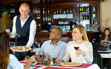 Friendly smiling waiter bringing ordered pizzas to guests of cozy italian restaurant