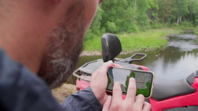 Slowmo tracking with close up of bearded man holding mobile phone and looking at digital map while standing in forest by the river. Quad bike parked before him
