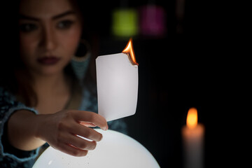 Portrait of Asian beautiful Gypsy fortune teller woman in dark room with illuminated light crystal ball and hold burning white card. Focused on hand.