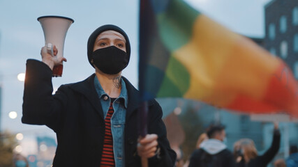 Young rebellious woman with face mask and megaphone holding rainbow flag in crowd. High quality...
