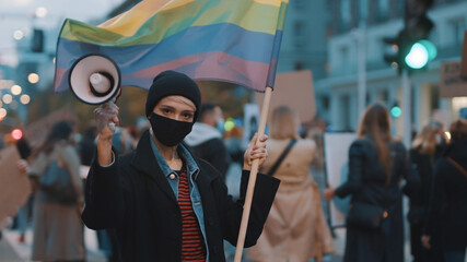 Young rebellious woman with face mask and megaphone holding rainbow flag in crowd. Protest against discrimination. High quality photo