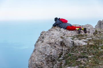 Girl lying on the ledge of the mountain looks into the abyss against the background of clouds and blurred outlines of the distant sea