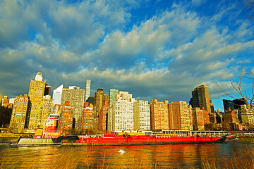 A view on Manhattan from Roosevelt Island in New York, USA. Waterfront buildings illuminated by sunrise under blue sky with cumulus clouds.