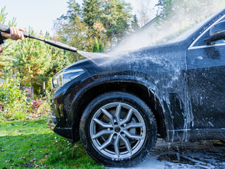 A high-pressure water jet sprays foam onto a black car in a backyard
