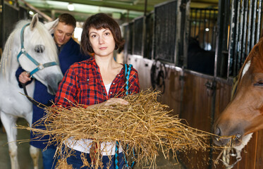 Female farm worker spreading armfuls of hay at horse stable