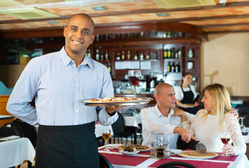 Smiling waiter holding serving tray with pizza at restaurant with customers his behind