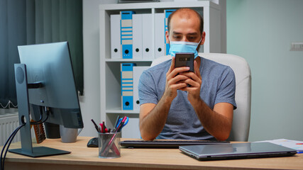 Office worker with protective mask using phone typing sitting on modern office during coronavirus. Freelancer working in new normal workplace chatting talking writing using mobile internet technology