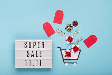 11.11 Super Sale text on white lightbox, shopping trolley, gift boxes and red tags on blue paper background.