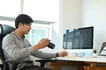 A young man photographer is checking previews on camera while sitting at his workspace at studio.