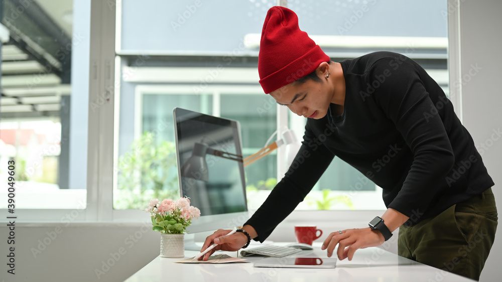 Poster concentrated young male designer standing at table with computer and examining paper with colour sam