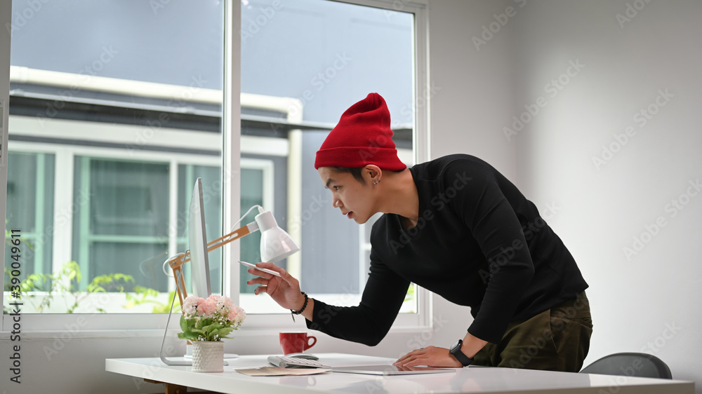 Poster A Graphic designer man in red wool hat standing at table with computer and examining paper with colour samples while working on project in office.