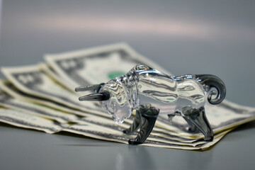 Figurine of a bull with banknotes on a gray background. Business concept, stock exchange, finance. Selective focus. US dollars are on top of each other and a glass bull standing above.