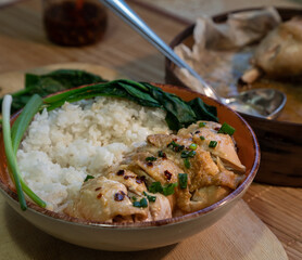 Chicken thigh sliced in Chinese or Asian Style served with steamed rice and greens on an orange plate. In the background is a wooden steamer with metal ladle. Hainanese chicken rice.
