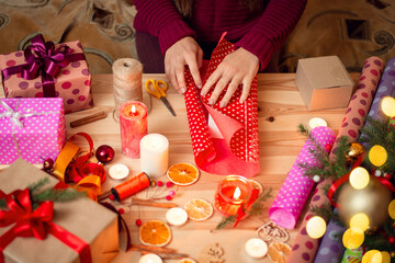 Hands of a woman in dark red sweater wrapping Christmas present on a wooden desk. Candles and festive decor, warm colours.