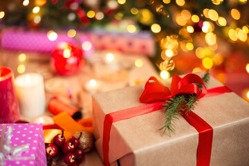 Gift box with red ribbon on the table near other presents. Bright lights from candles and christmas tree on the background.