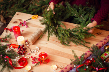 Light wooden table with fir-tree branches and festive decoration. Christmas wreath in hands of a woman making it.