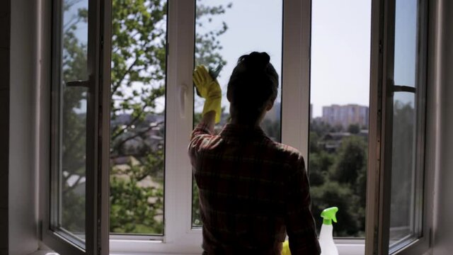 Housewife in protective yellow gloves cleaning window with spray and rag