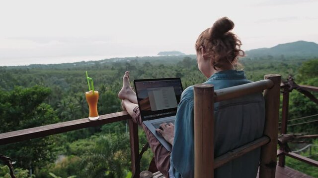 Freelancer woman sits with a laptop on a high bar stool. Amazing sunset views and dense jungle by the blue sea.