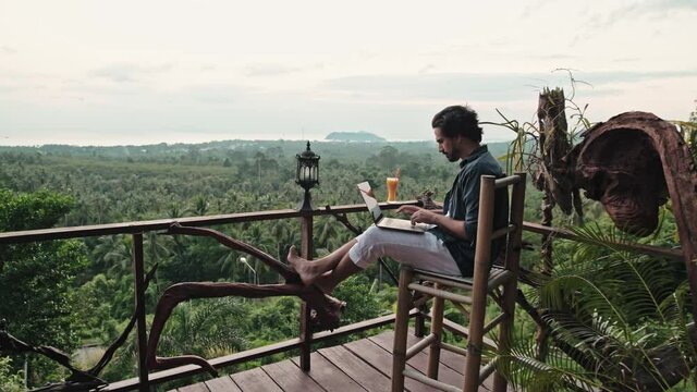 A young, handsome man with a beard works on a laptop in a cafe overlooking an incredible sunset.