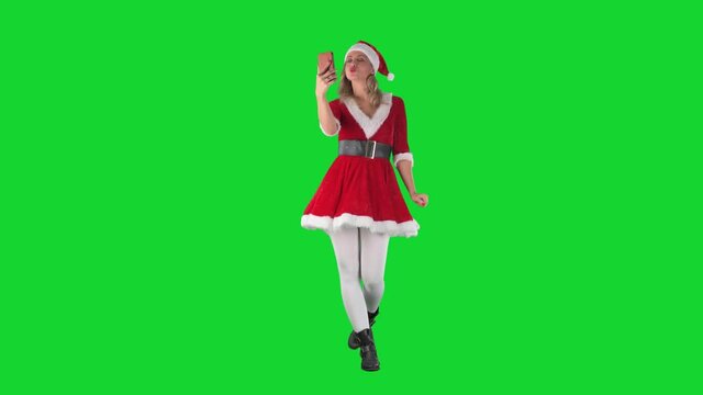 Modern Santa Claus girl walking and taking selfie photos kissing and waving at cellphone. Full body length on green screen background. 