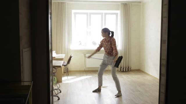 Positive woman dancing and singing with broom in room during housework and having fun