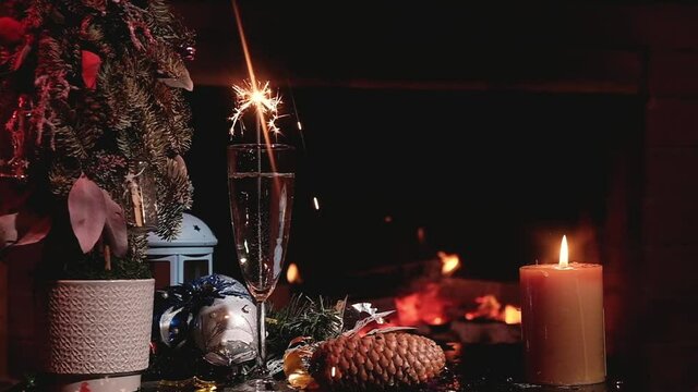 Christmas drink champagne in glasses with burning Sparkler on background festive fireplace with gift box, artificial Christmas tree and candle burning. Sparkling wine on festive table