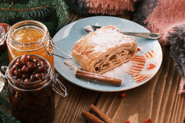 Closeup of a strudel on a Christmas plate near cinnamon sticks and a jam jars. Christmas breakfast on a wooden table
