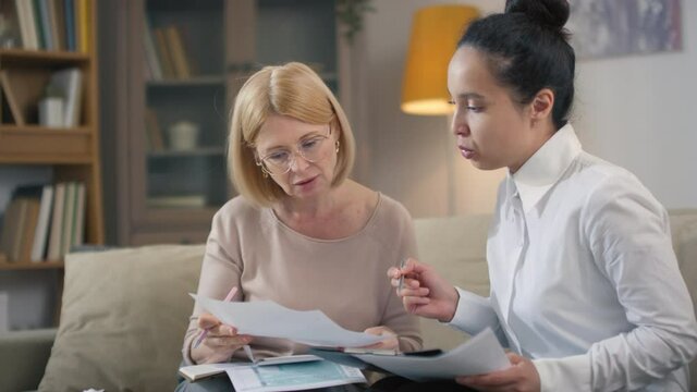 Medium closeup of young mixed race financial consultant in white shirt explaining middle-aged woman basics of financial literacy and planning while sitting at home office going through papers