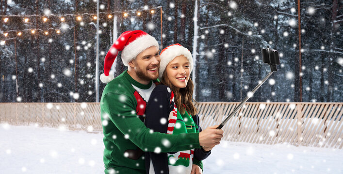 people, technology and holidays concept - happy couple in santa hats and ugly sweaters taking picture with smartphone on selfie stick over skating rink and snow on background
