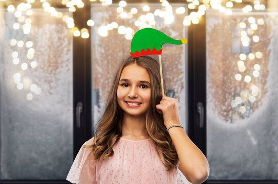 christmas, holidays and photo booth concept - happy smiling teenage girl with santa helper hat party accessory over garland lights on snowy window background