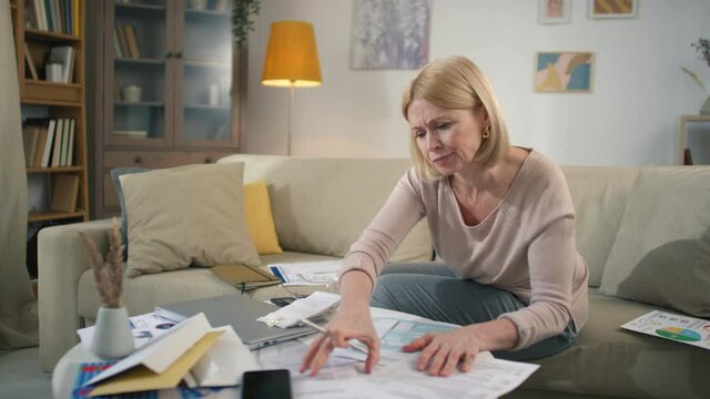 Stressed middle aged woman looking through piles of papers on messy table while analyzing finances getting upset and frustrated