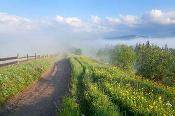 Mountain road with a wooden fence and a bright meadow, foggy mountains on the horizon.