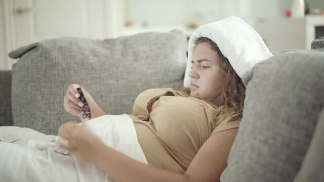 Concentrated young plump woman choosing pills lying on couch with towel on head. Side view portrait of ill Caucasian plus-size lady having cold and flu, fever, and high temperature.