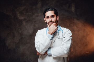 Bearded and cheerful doctor in labcoat with stethoscope on his neck poses with hand under his chin in dark background.