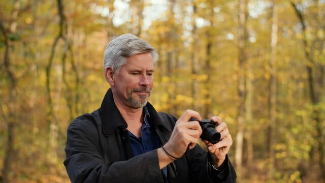 Mature middle-aged white man taking pictures in a scenic forest in late autumn. Handsome male photographer travels outdoors with a small camera. Beautiful nature background during fall. Happy tourist.