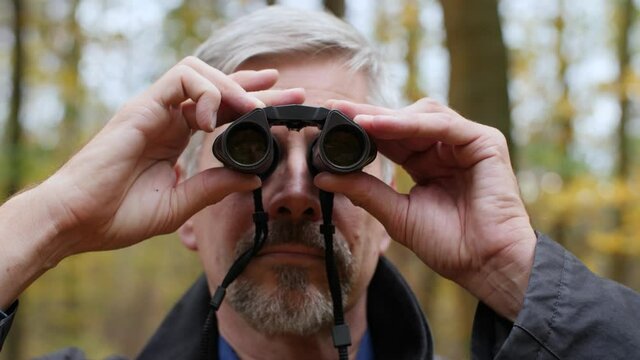 Mature middle-aged white man using binoculars in a scenic forest in late autumn. Handsome guy watches birds outdoors and enjoys his hobby. Beautiful nature background during fall. Happy lifestyle.
